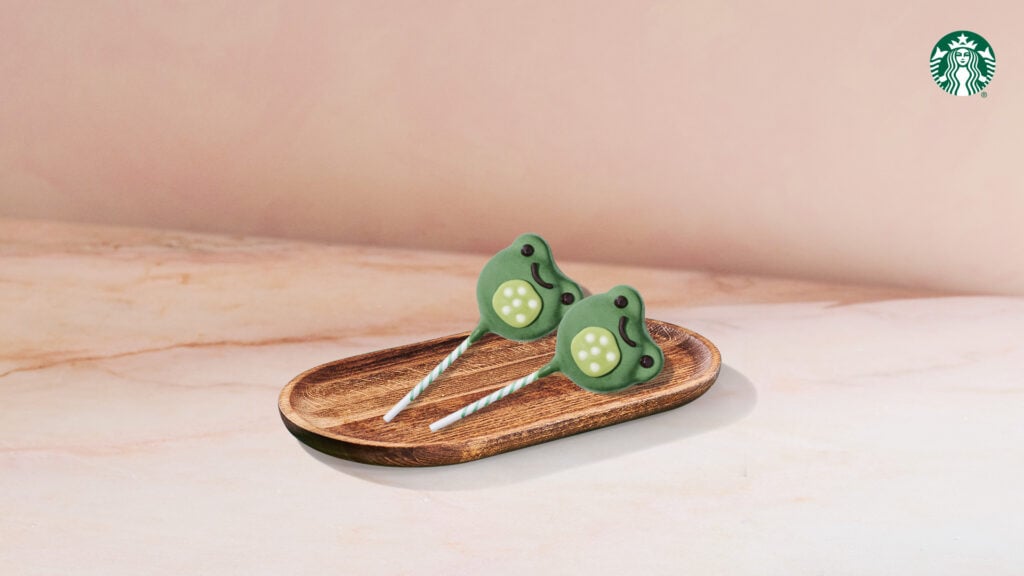 Two frog-shaped cake pops with green coating and decorative features are placed on a wooden tray against a soft background.