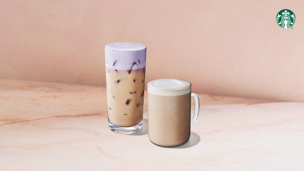 Two coffee beverages are displayed on a light marble surface. On the left, a tall glass holds iced coffee topped with a purple foam layer. On the right, a short glass mug contains a creamy, warm coffee drink with a smooth surface.
