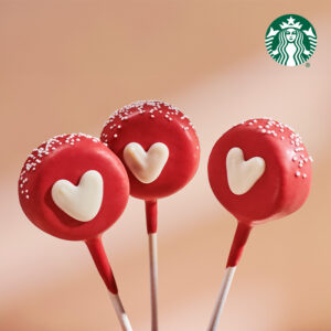 Three red cake pops with white heart designs and sprinkles on top, presented on sticks against a soft background.