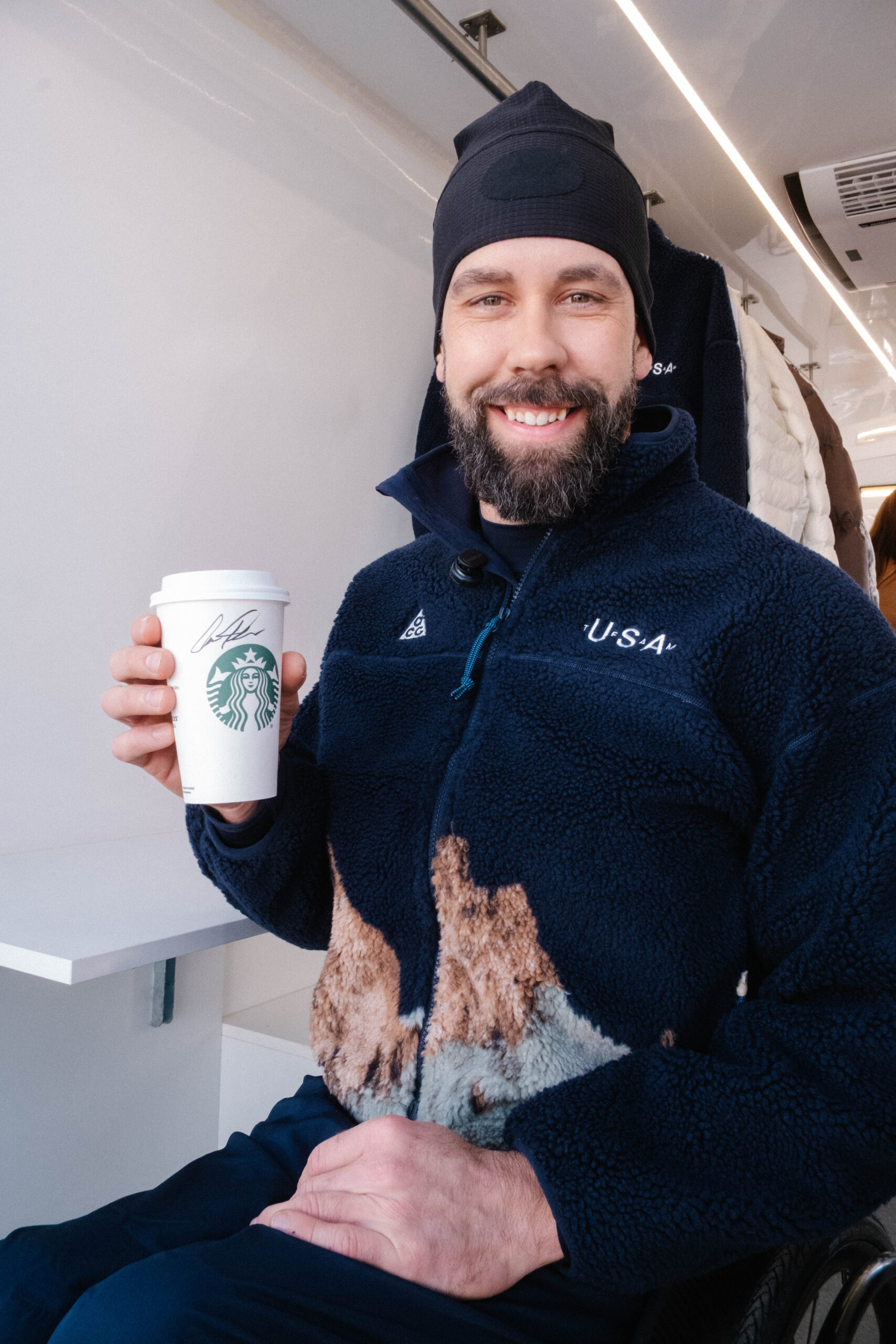 A man with a beard smiles while holding a Starbucks cup. He wears a navy fleece jacket with a design and a black beanie.