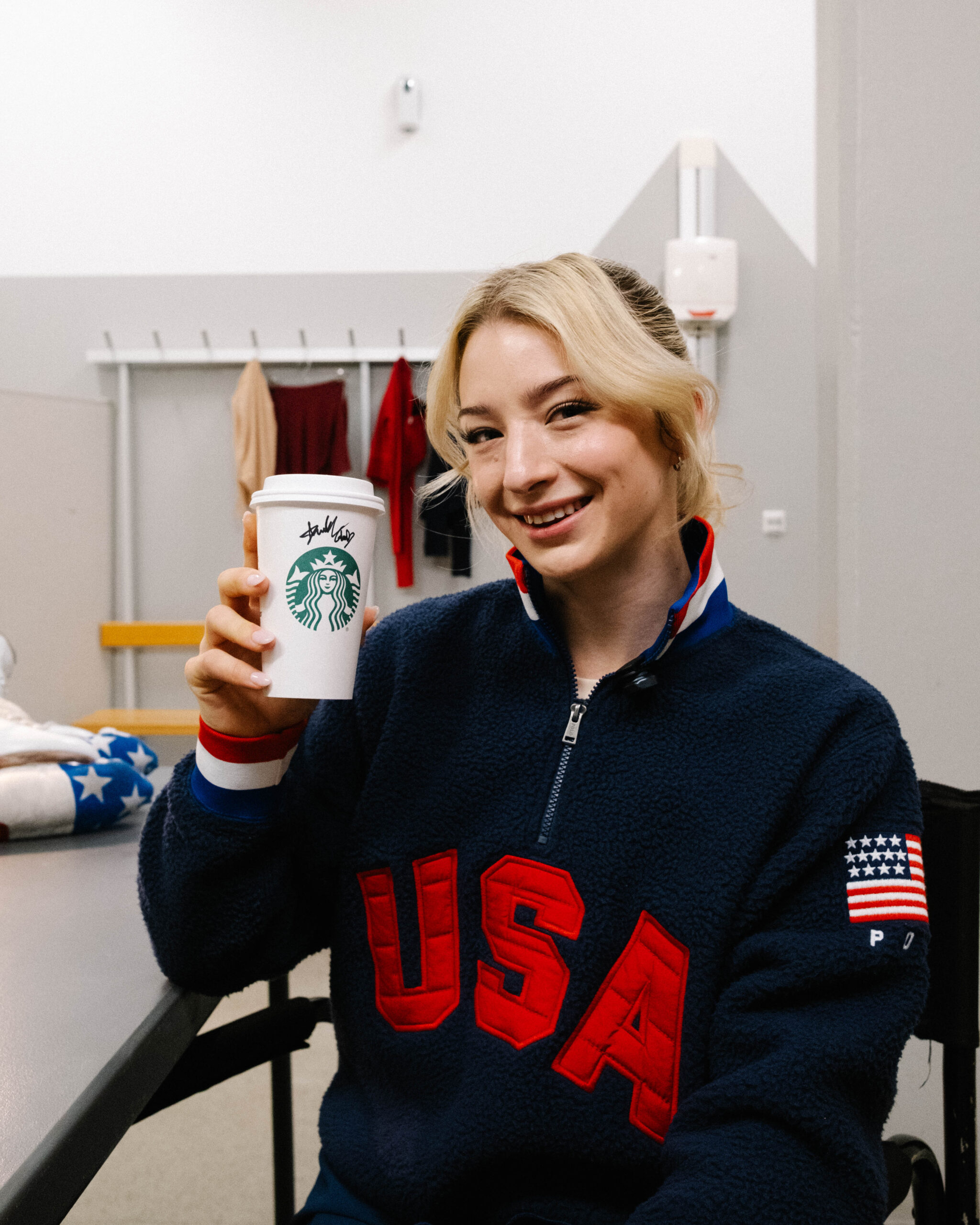 A person in a navy fleece jacket with "USA" in red is smiling and holding a Starbucks cup. The background features clothing on hooks.