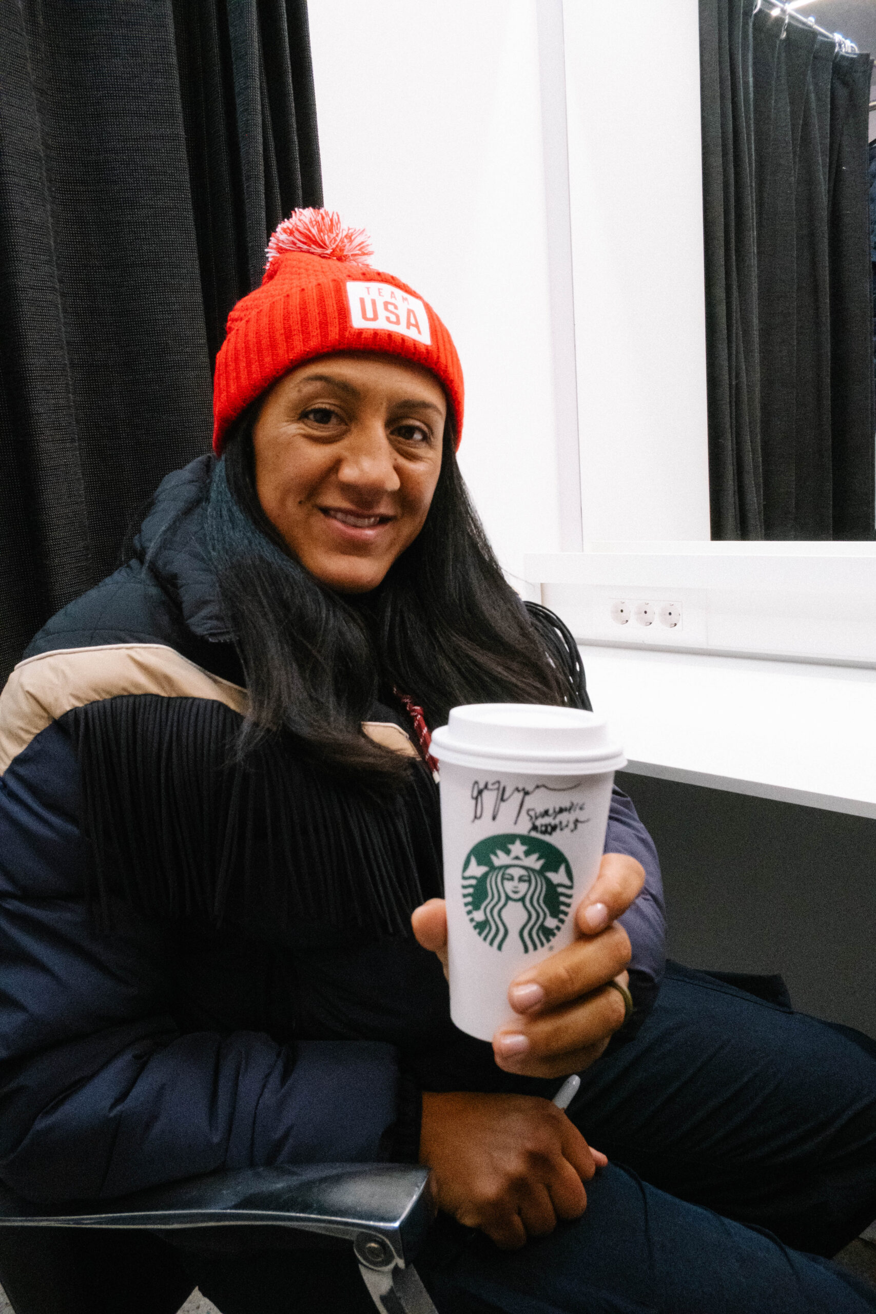 A person wearing a red "Team USA" beanie and a dark jacket holds a Starbucks cup with a signature on it, sitting in front of a mirror.