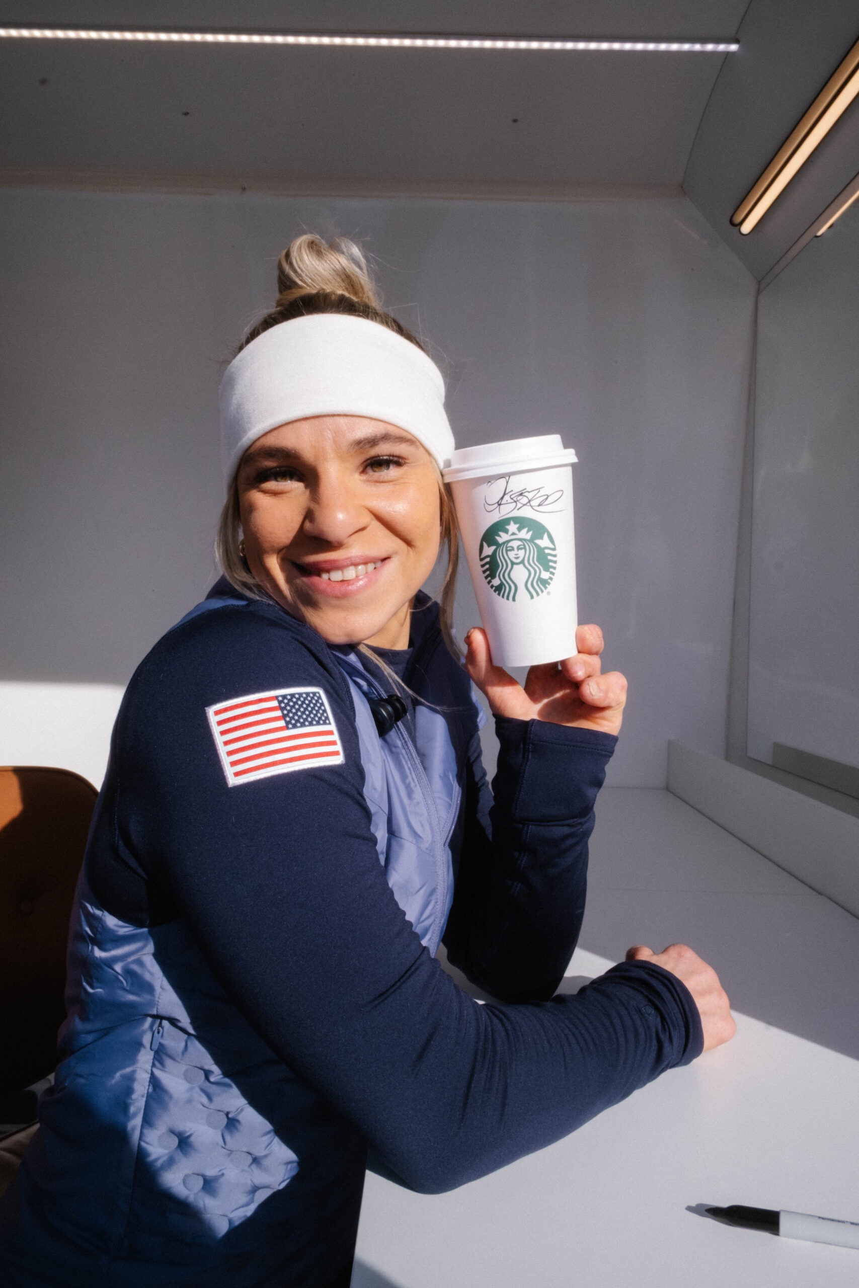 A smiling woman holds a Starbucks cup, wearing a white headband and a navy blue jacket with a light blue vest. A pen is on the table.