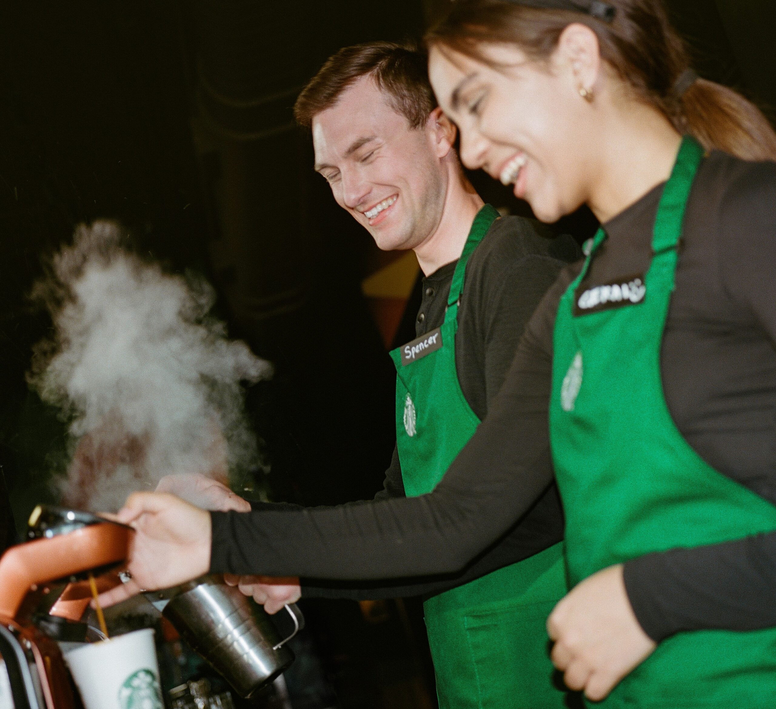 Two baristas in green aprons are smiling while preparing drinks. Steam rises from a kettle as one pours into a cup.