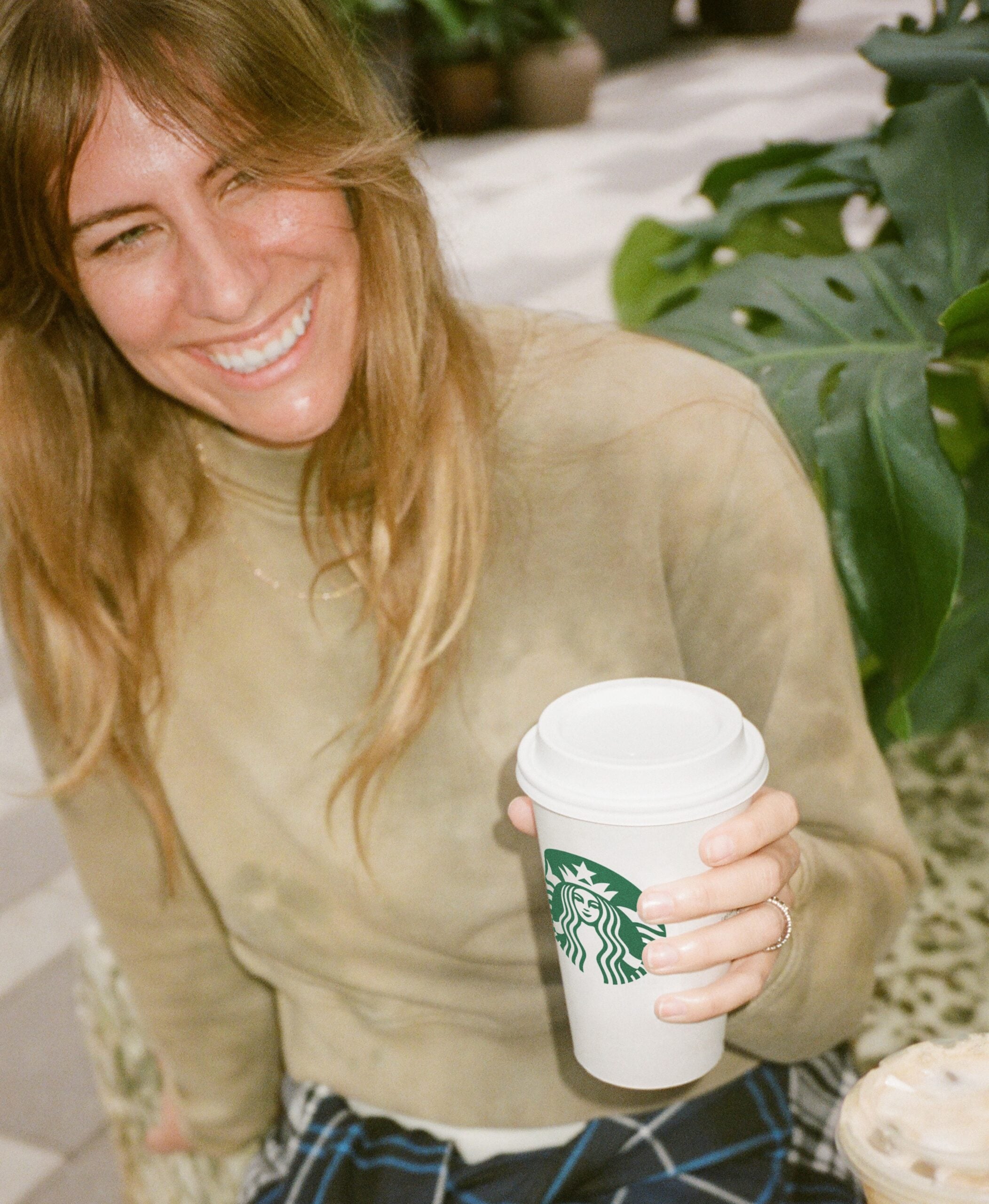 A smiling woman with long, wavy hair holds a Starbucks coffee cup. She wears a light brown top and is seated among greenery.