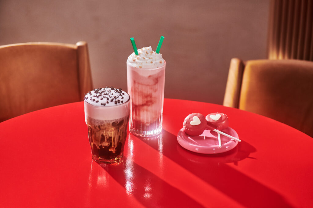 Two colorful drinks sit on a bright red table. On the left, a glass of iced coffee topped with whipped cream and chocolate sprinkles. On the right, a tall glass of a creamy beverage with whipped cream and green straws. Nearby, a pink plate holds two heart-shaped treats on sticks.