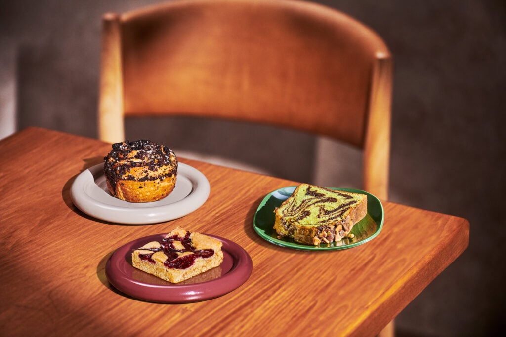 A wooden table features three desserts: a chocolate swirl muffin on a gray plate, a jam-topped square on a maroon plate, and a green marbled cake on a green plate. A wooden chair is partially visible in the background.