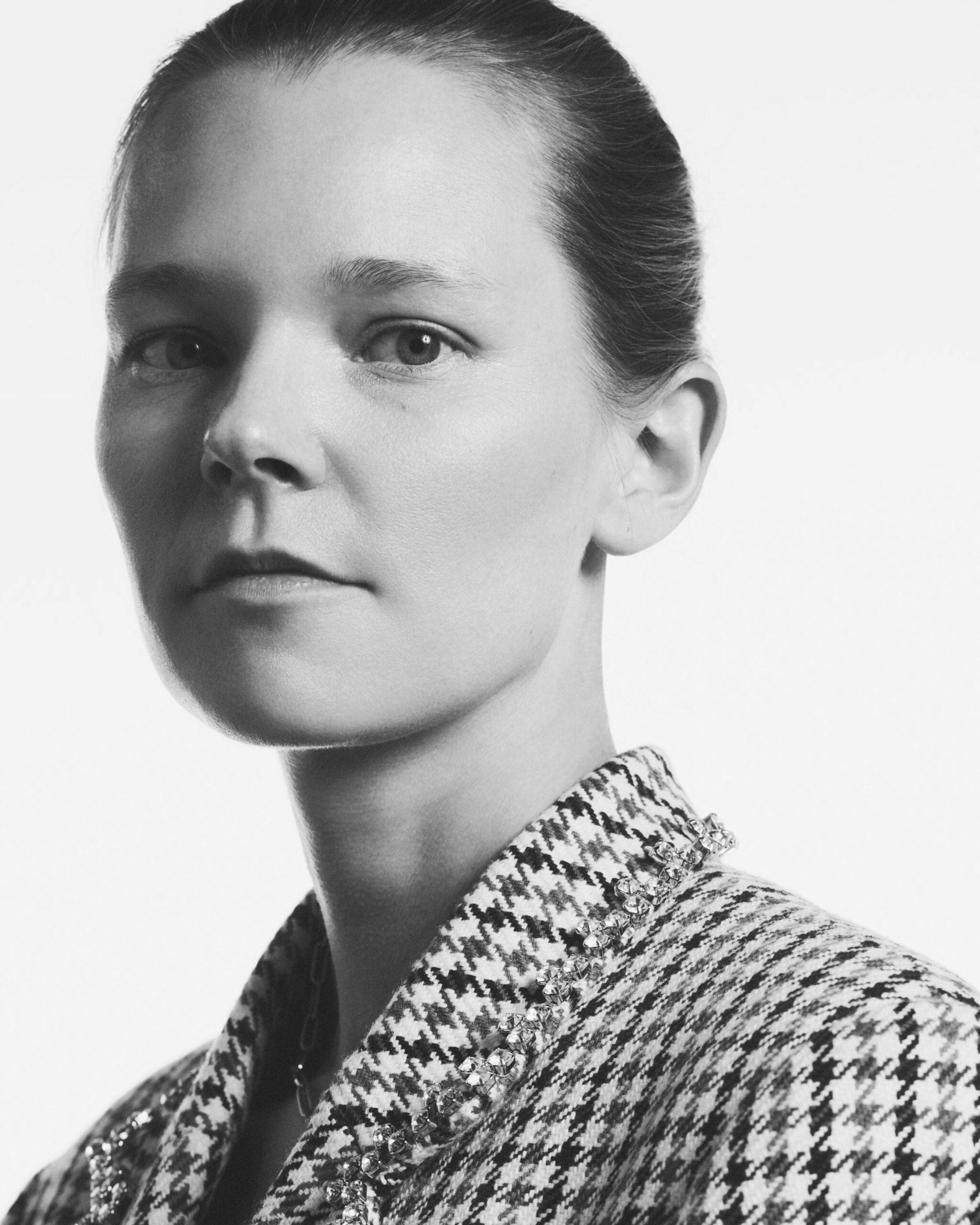 A close-up portrait of a woman with a sleek hairstyle, wearing a patterned jacket with embellishments. The background is white, emphasizing her features.