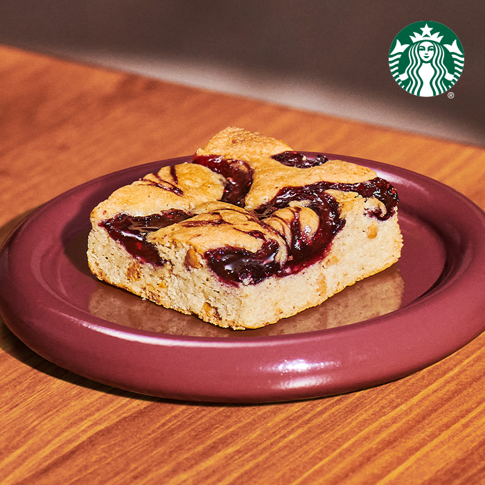 A square piece of baked dessert with a golden-brown top, featuring swirls of dark berry jam, served on a maroon plate.