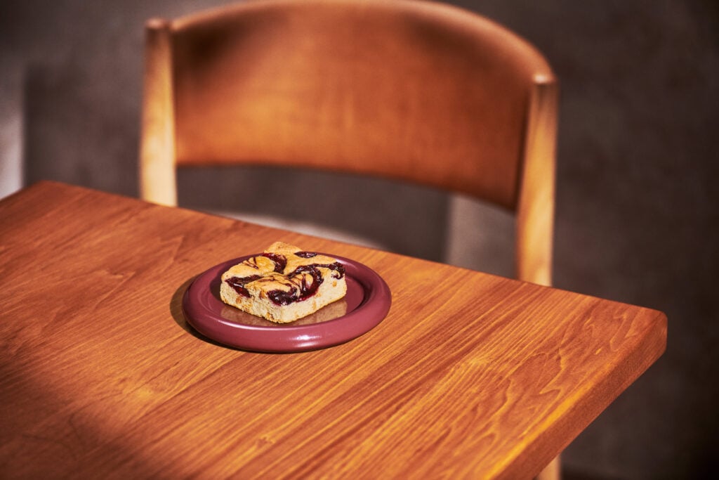 A slice of dessert with a fruit topping sits on a round, maroon plate on a wooden table. A wooden chair is partially visible in the background.