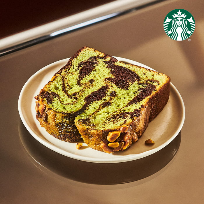 A slice of marbled green and chocolate bread is presented on a white plate, showcasing a swirl pattern and topped with nuts.