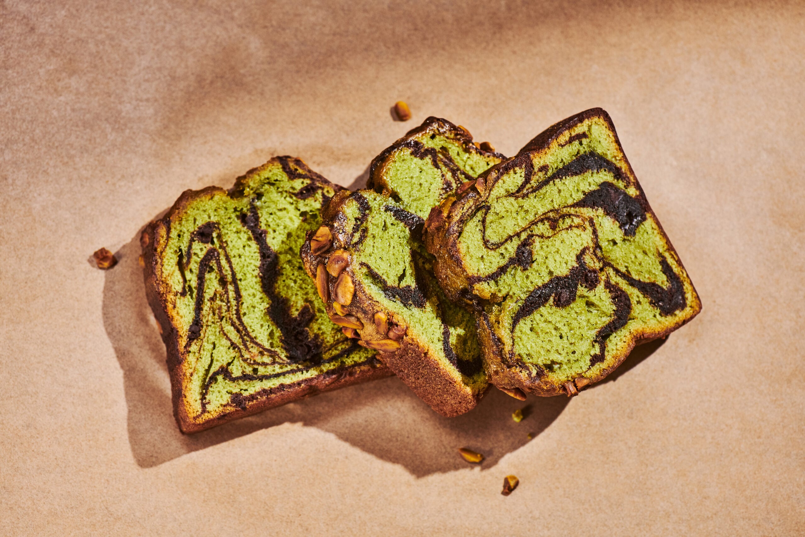 Three slices of green and chocolate swirl bread are arranged on a brown paper surface, showcasing a marbled pattern and nutty topping.
