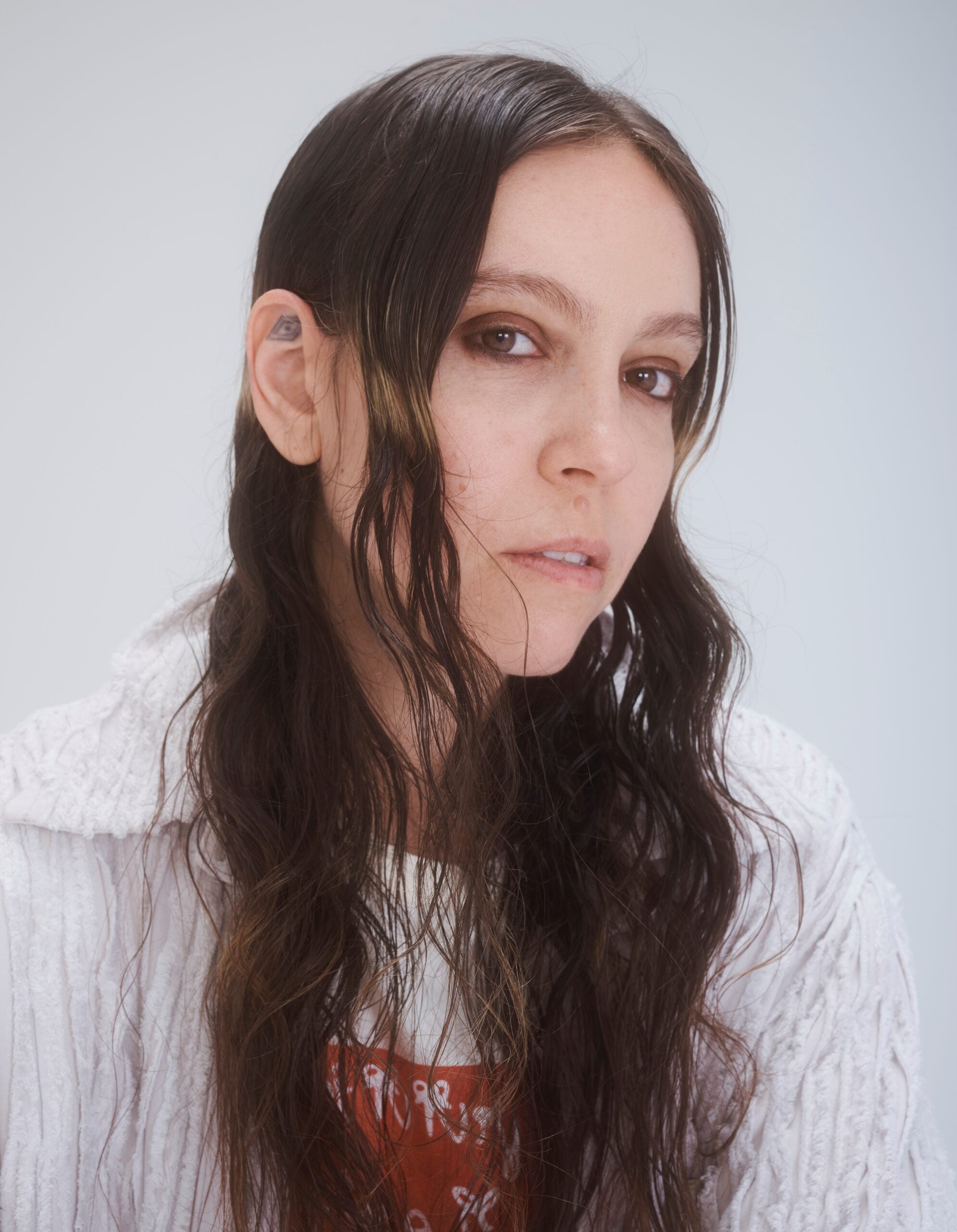 A person with long, wavy hair is looking directly at the camera. They wear a white textured sweater over a graphic t-shirt. The background is light and neutral.