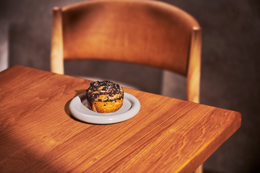 A cinnamon roll topped with dark crumbs sits on a light gray plate, placed on a wooden table. A wooden chair is visible in the background.