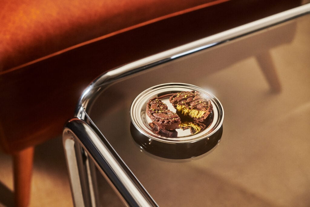 A shiny silver plate holds two chocolate cookies, one cut in half to reveal a bright yellow filling, topped with green sprinkles. The plate rests on a reflective glass table.
