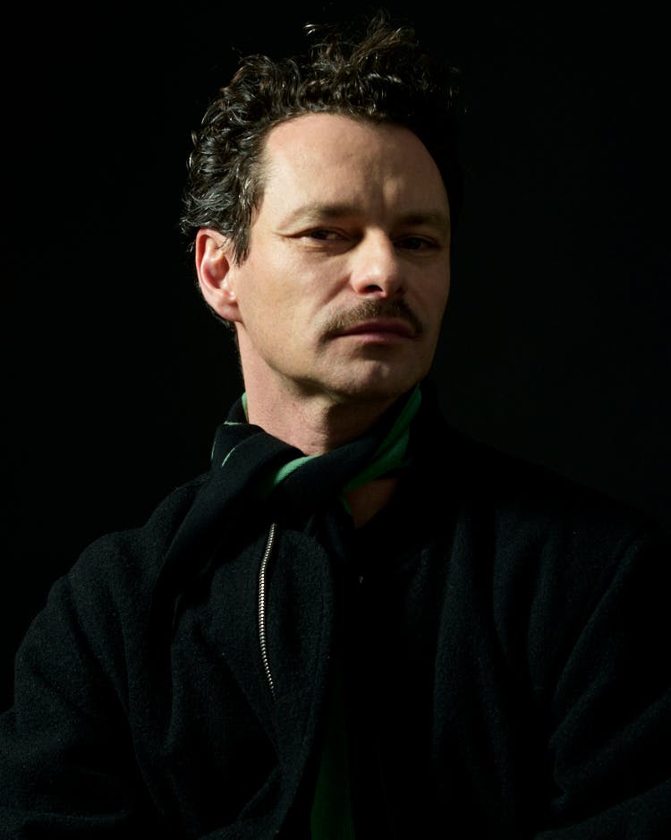 A man with curly hair and a mustache wears a black coat and a green scarf, posing against a dark background.