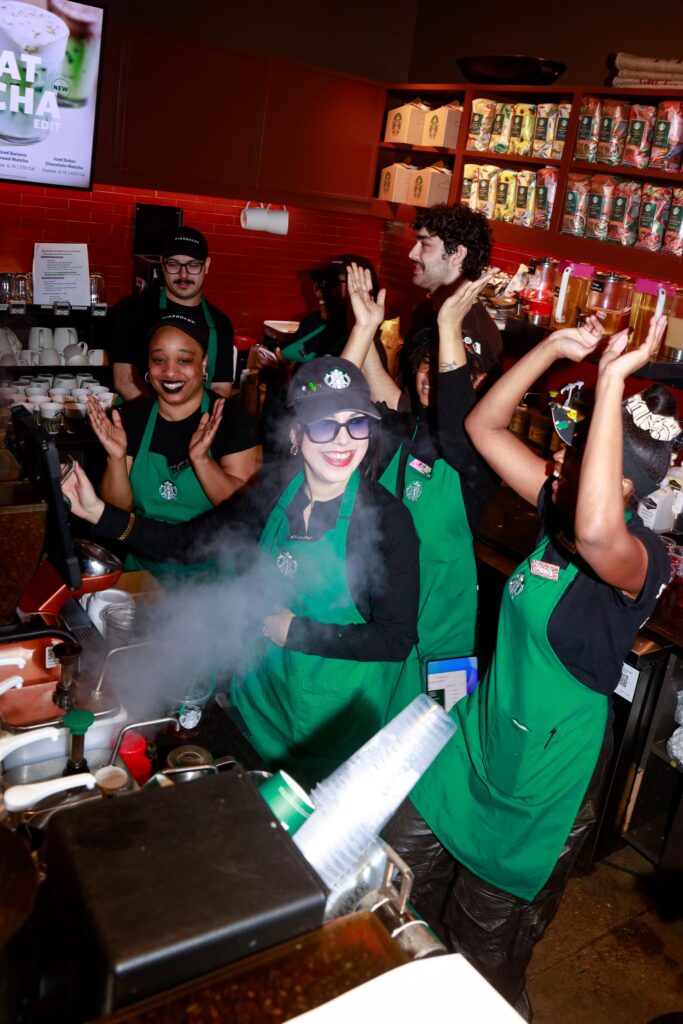 A lively Starbucks scene with baristas in green aprons celebrating. Steam rises from the coffee machine as they cheer and dance.