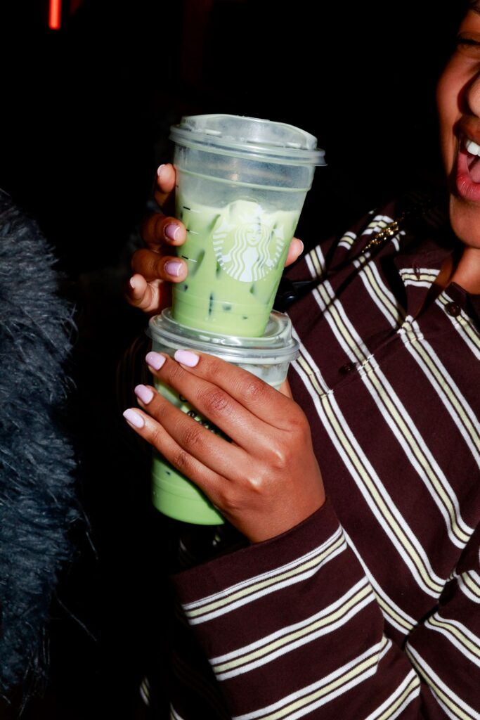 A person holds two iced green beverages in clear cups, featuring a logo. Their nails are painted in a light pink shade.