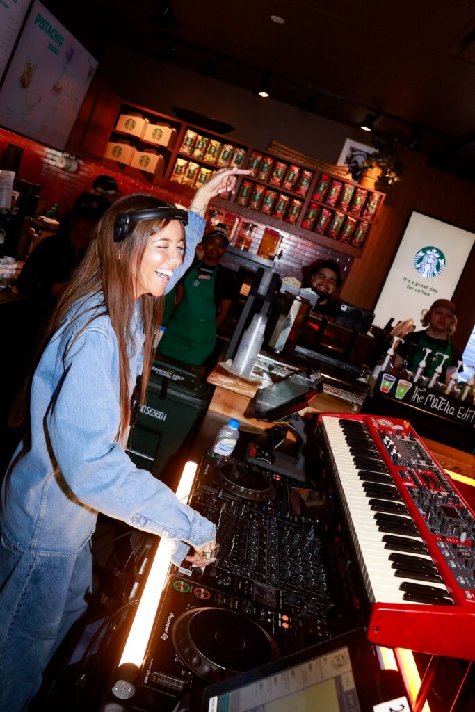 A DJ is mixing music at a Starbucks, smiling and wearing headphones. Behind her, baristas are engaged and enjoying the atmosphere.