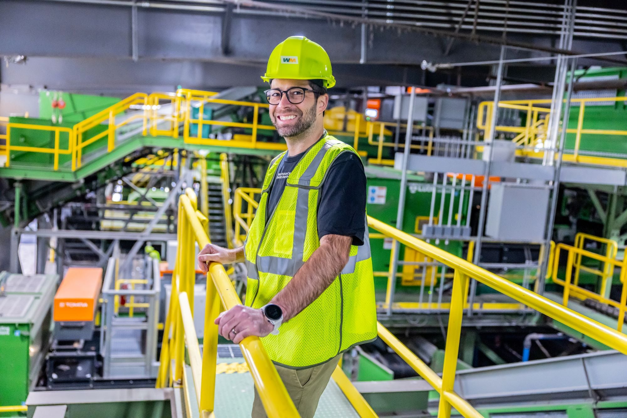 A person wearing a yellow hard hat and a reflective safety vest stands on a railing in an industrial facility. The background features green machinery and yellow safety railings, indicating a recycling or waste management environment.
