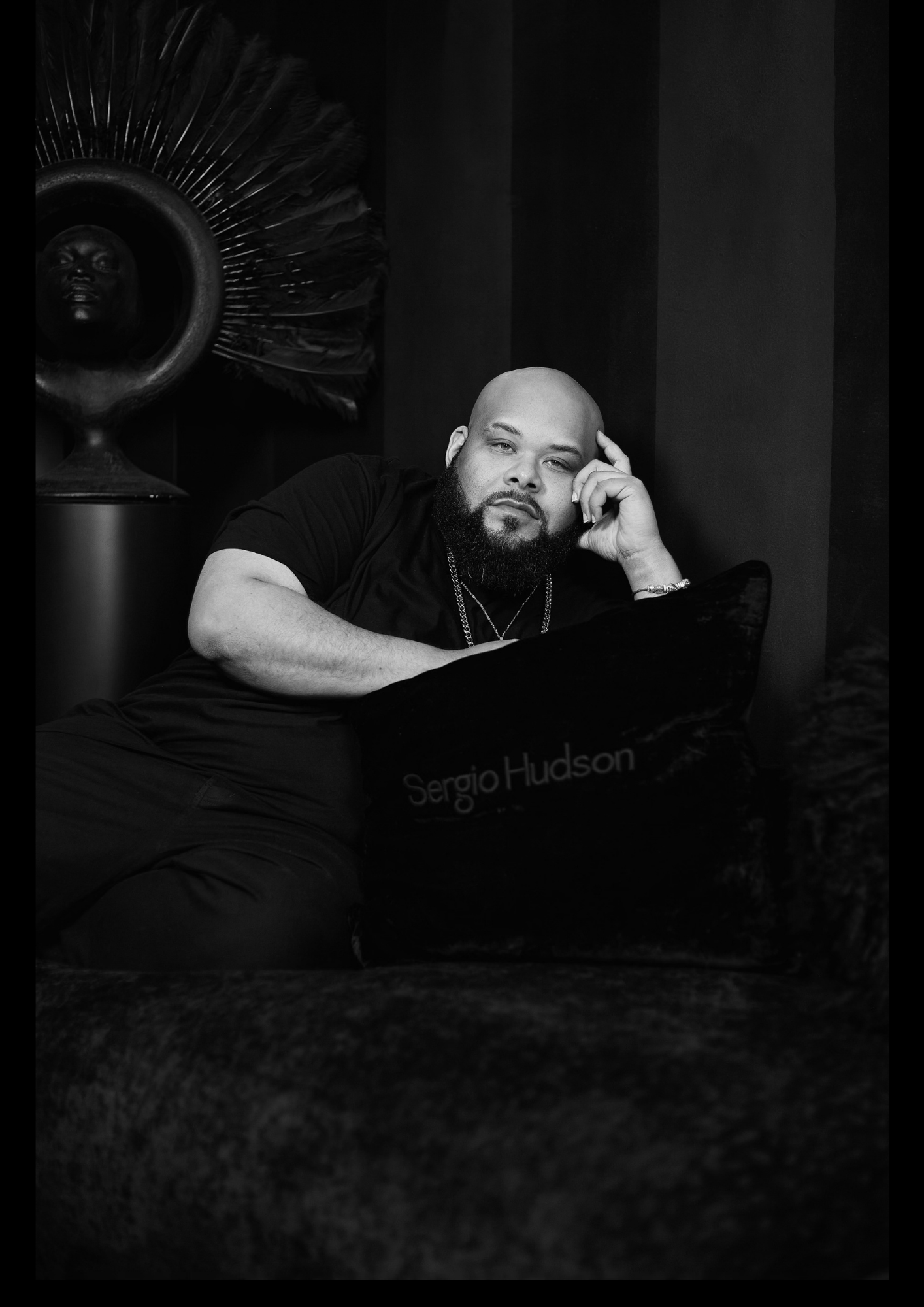 A man with a beard sits thoughtfully on a plush couch, wearing a black shirt. A decorative piece is visible in the background.