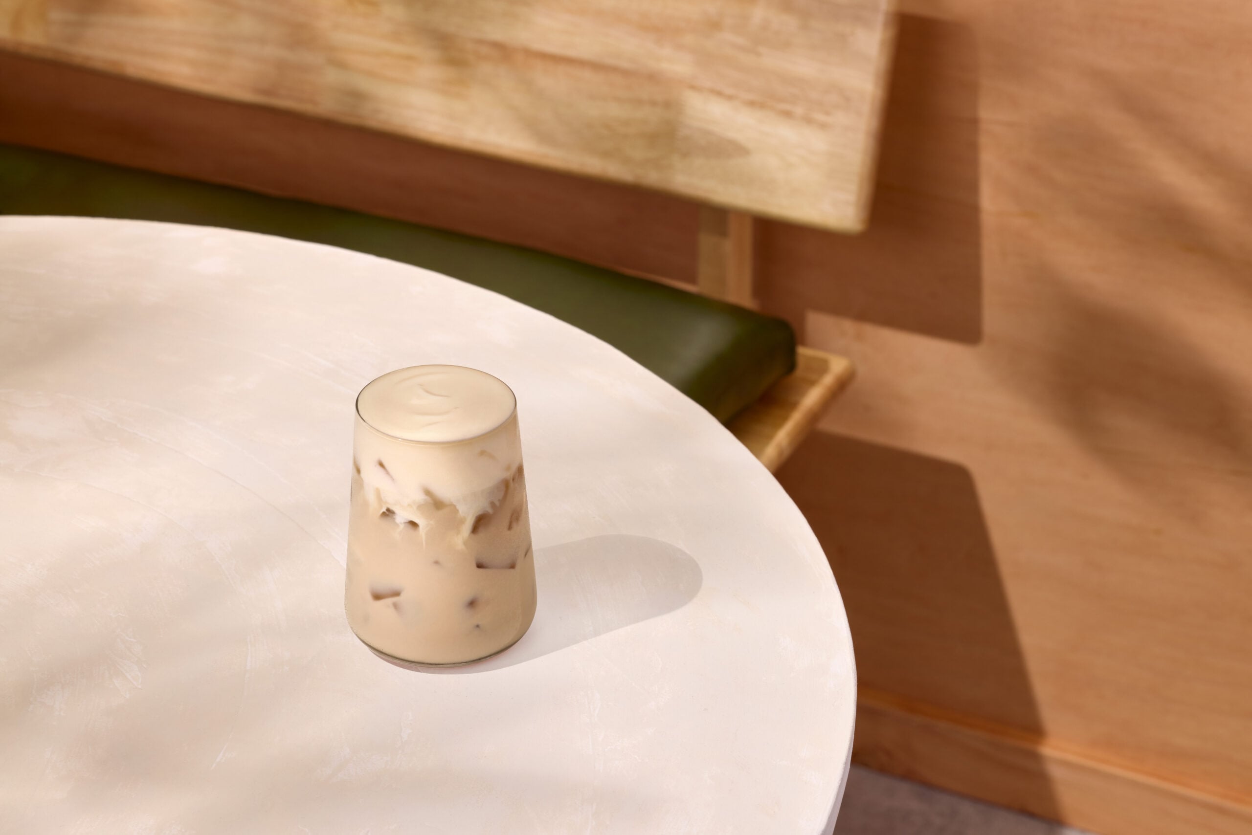 A glass of iced coffee sits on a round white table. The drink has a creamy top and visible ice cubes. A green bench is in the background.