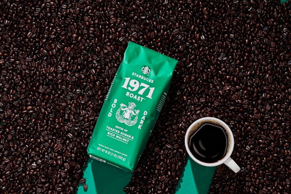 A green bag of Starbucks 1971 Roast coffee sits among dark roasted coffee beans, with a white cup of black coffee nearby.