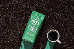 A green bag of Starbucks 1971 Roast coffee sits among dark roasted coffee beans, with a white cup of black coffee nearby.