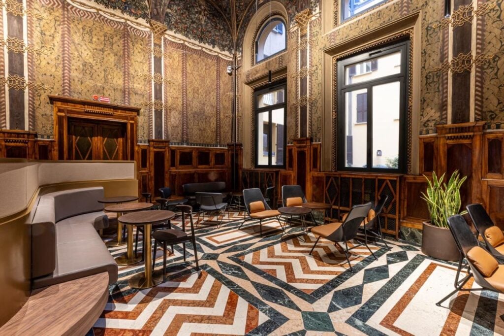 A stylish interior featuring ornate wallpaper, wooden paneling, and a patterned marble floor. Seating includes modern chairs and small tables.