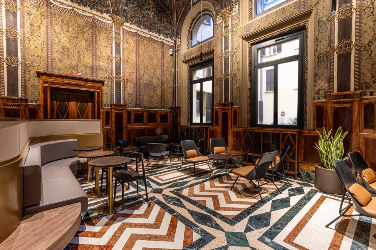 A stylish interior featuring ornate wallpaper, wooden paneling, and a patterned marble floor. Seating includes modern chairs and small tables.