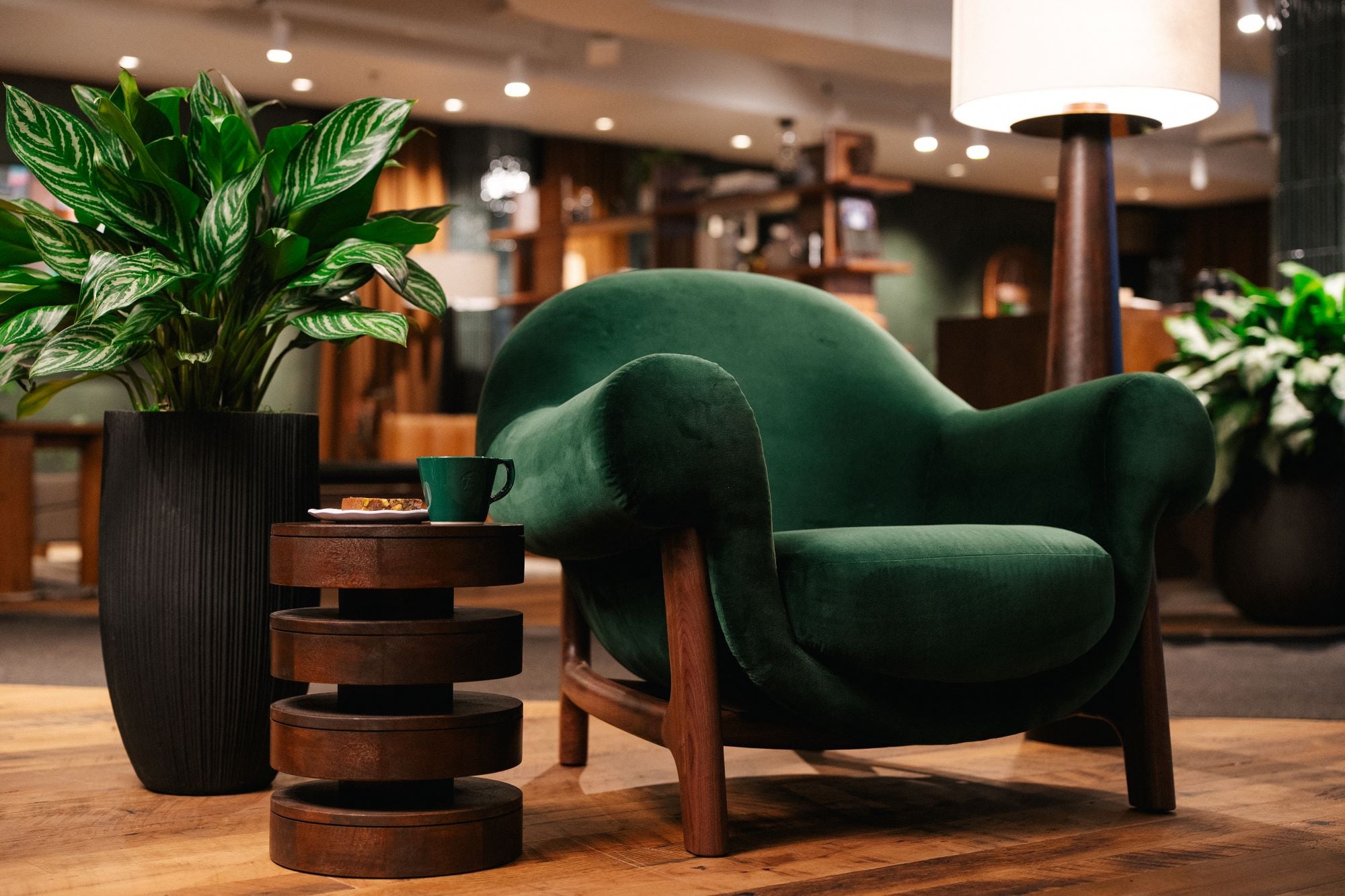 A plush green armchair with rounded edges sits beside a dark wooden side table. A small green cup and a dessert are on the table, next to a tall, textured black planter filled with leafy plants. The background features warm lighting and wooden decor elements.