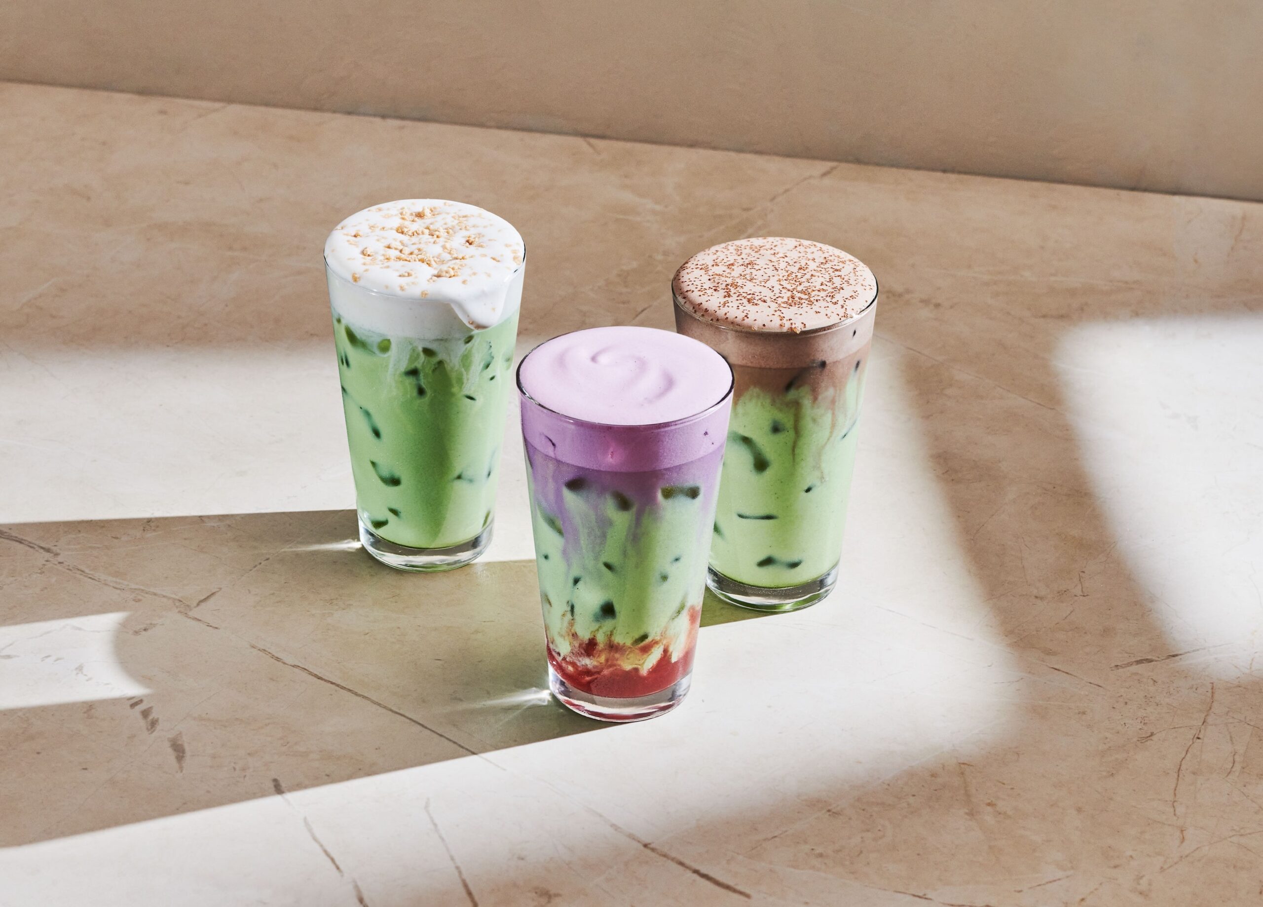 Three colorful drinks are displayed on a light marble surface. The left glass features a green beverage topped with whipped cream and crushed nuts. The center glass has a purple layer above a green base, while the right glass contains a brown drink with a frothy top dusted with cocoa powder.