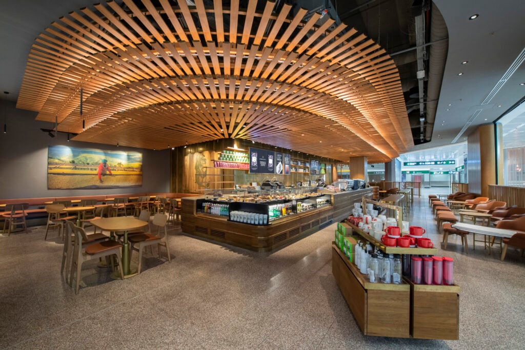 A modern café interior features a wooden slatted ceiling, a service counter with various beverages and pastries, and seating areas with round tables and chairs. The design is warm and inviting, with a mix of natural wood tones and soft lighting.
