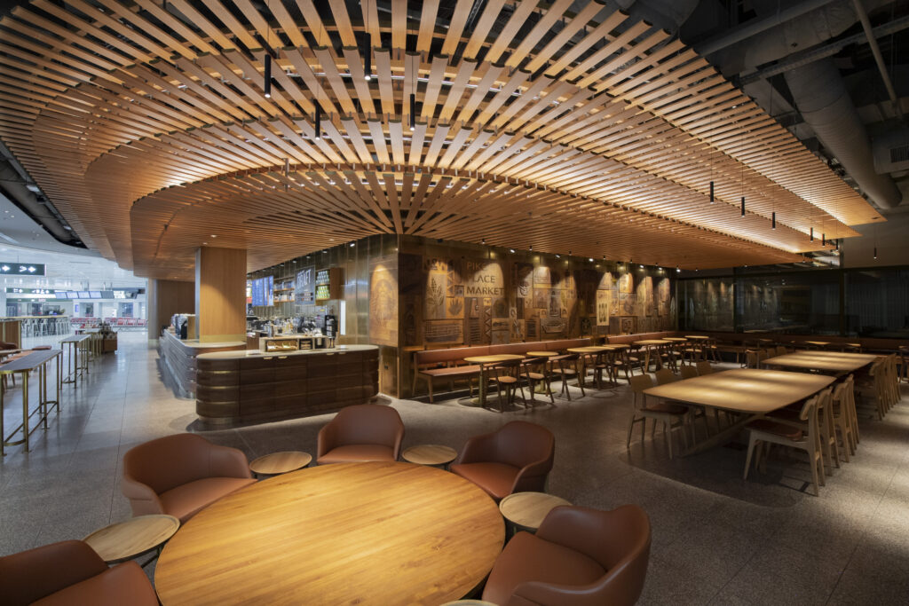 A modern café interior featuring wooden slatted ceilings, a bar area, and various seating arrangements, including round tables and cozy chairs. The walls are adorned with artistic engravings, creating a warm and inviting atmosphere.