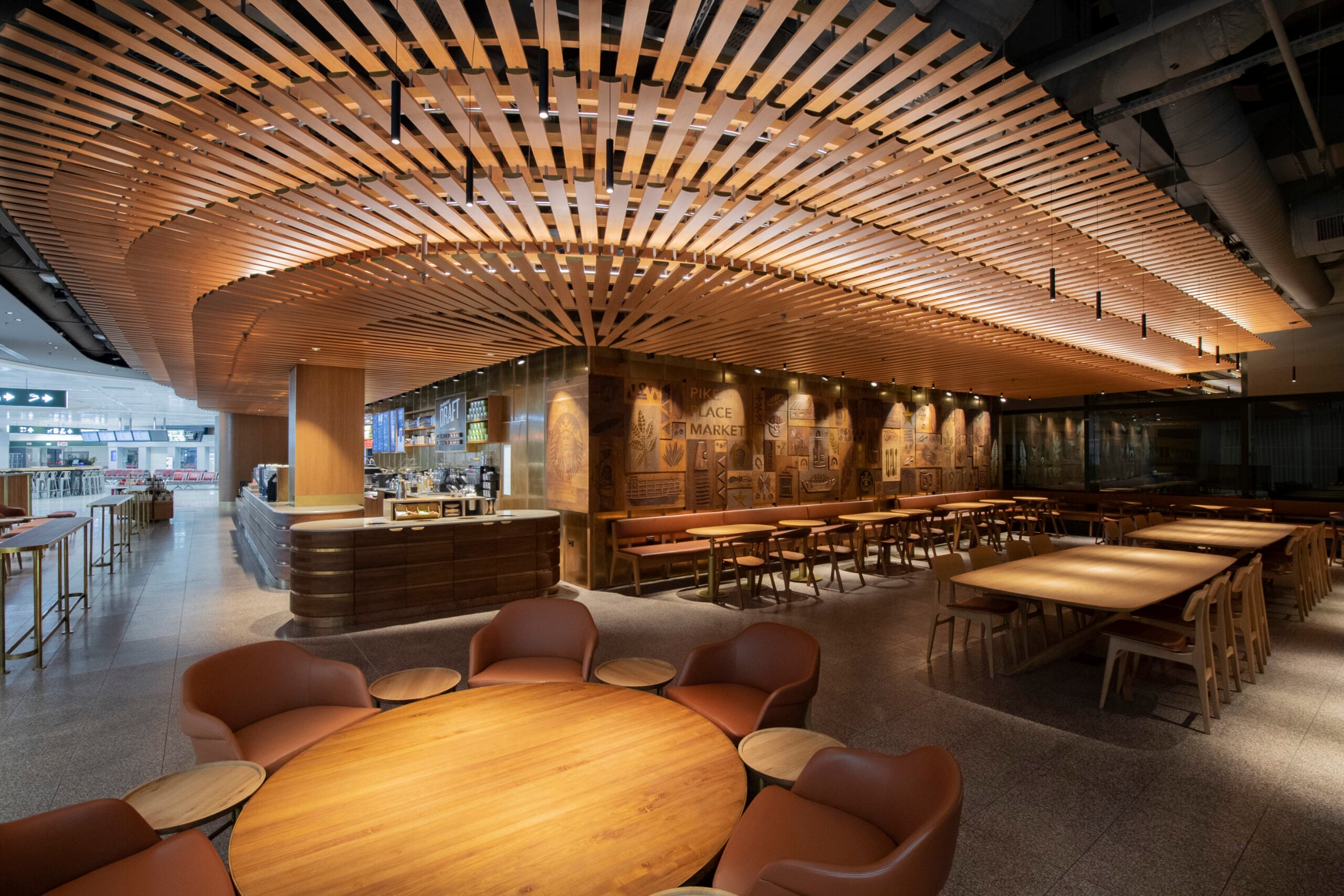 A modern café interior featuring wooden slatted ceilings, a bar area, and various seating arrangements, including round tables and cozy chairs. The walls are adorned with artistic engravings, creating a warm and inviting atmosphere.