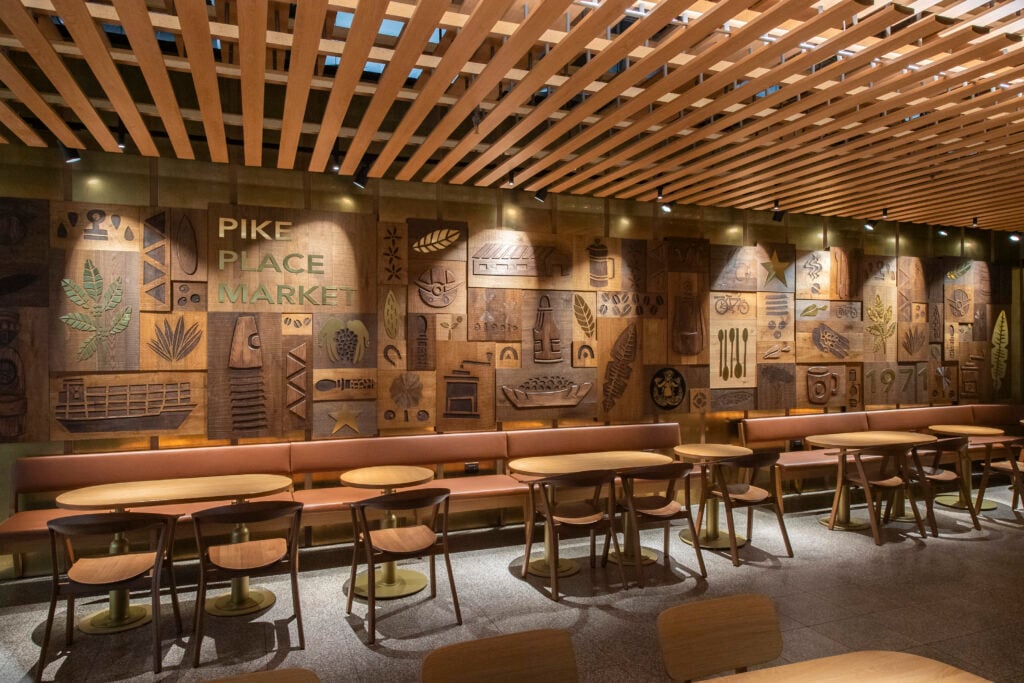 A cozy café interior featuring wooden paneling with carved designs, including leaves and various market-related motifs. There are round tables and chairs, with a long bench along the wall. The lighting is warm and inviting.