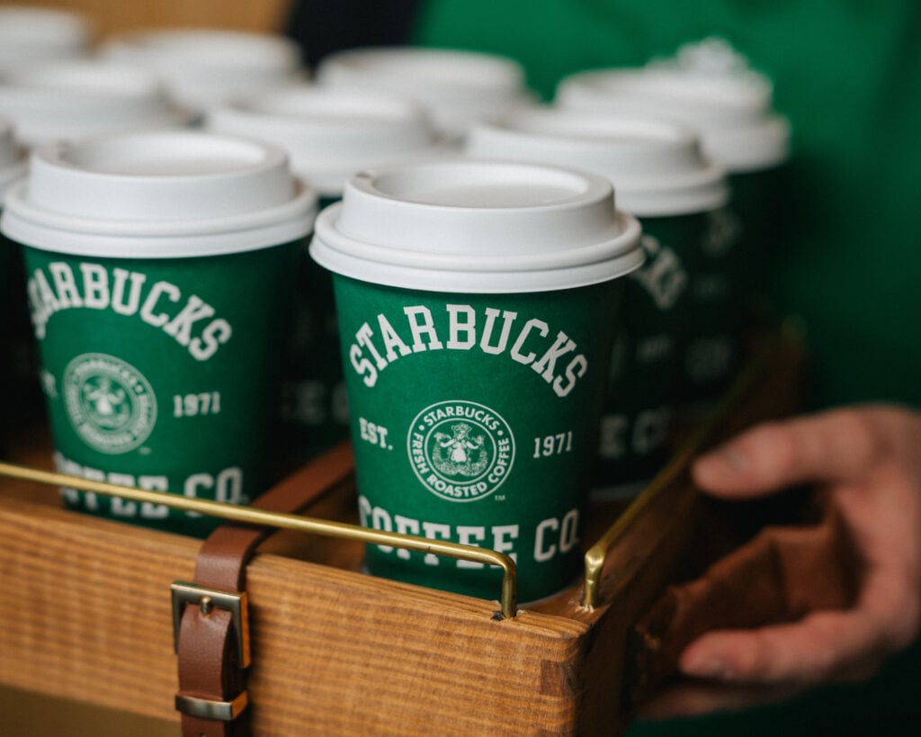 A wooden tray holds several green Starbucks coffee cups with white lids, featuring the brand's logo and established date.