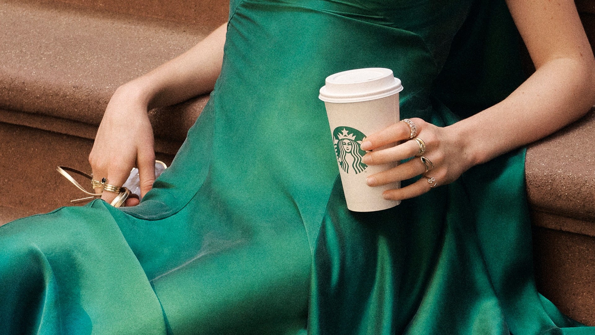 A hand holds a Starbucks coffee cup while wearing multiple rings. The person is dressed in a flowing green dress, seated on a stone surface.