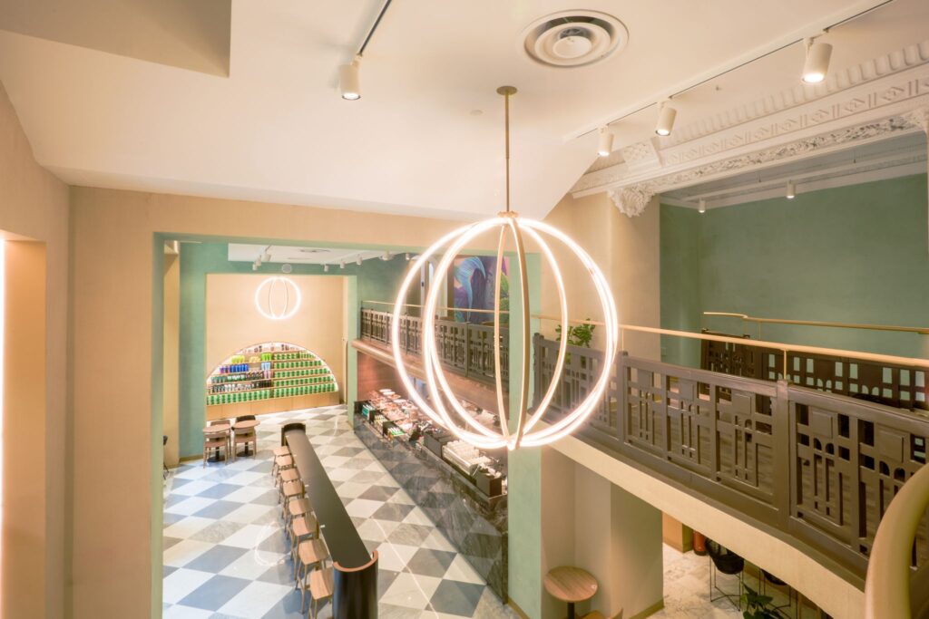 A modern café interior featuring a large circular pendant light, a long black counter, and a patterned floor. Shelves display colorful products.