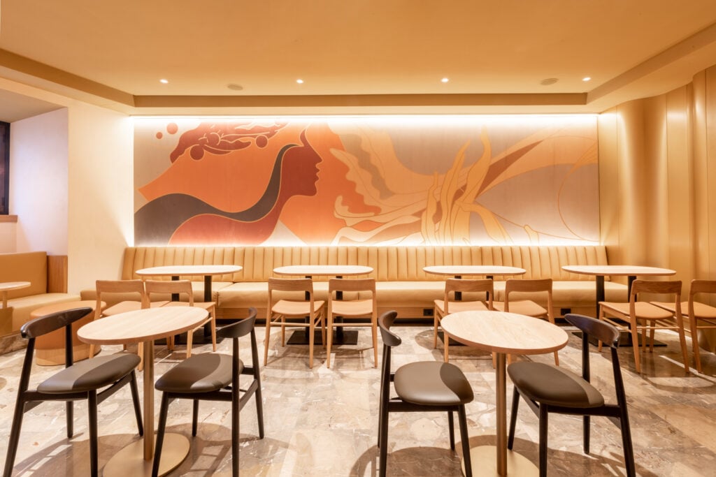 A cozy café interior featuring light wood tables and black chairs. A large, warm-toned mural adorns the wall, complemented by soft lighting.