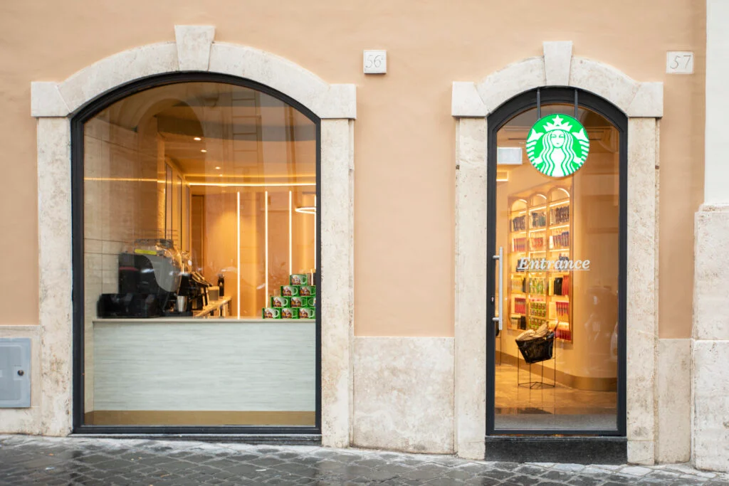 A storefront featuring two arched windows. The left window displays a coffee preparation area with equipment and stacked items. The right window has a door marked "Entrance" and showcases colorful products inside, along with the Starbucks logo above. The building has a light-colored facade with stone detailing.