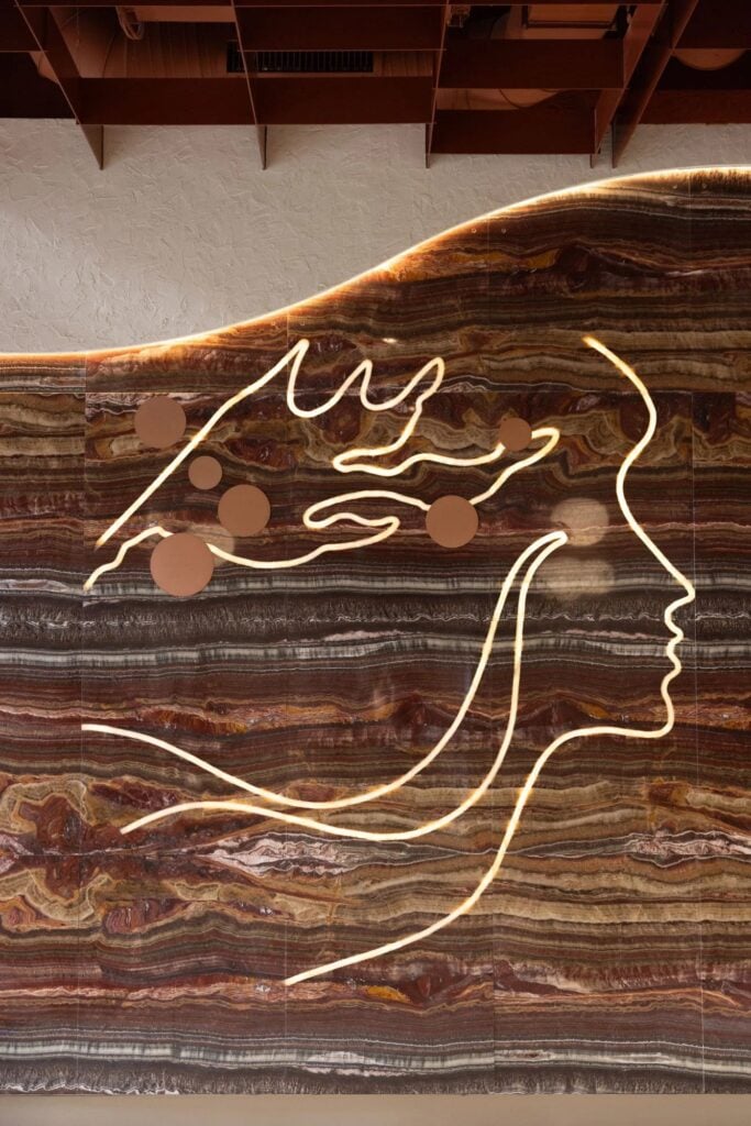 A textured wall features a flowing design of a face outlined in light, complemented by circular shapes against a rich, layered stone background.