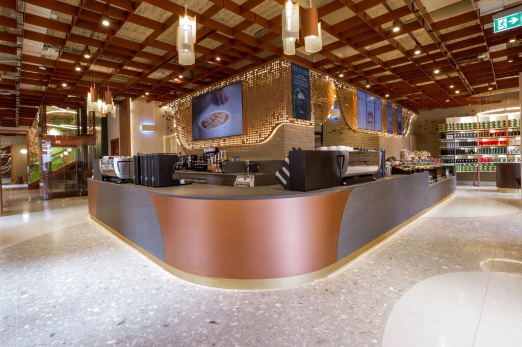 A modern coffee shop interior featuring a curved counter with a mix of materials, decorative lighting, and shelves stocked with products.
