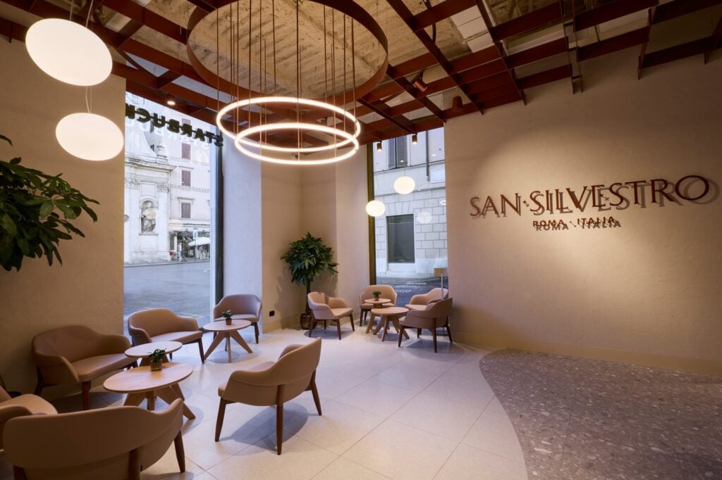 A modern interior featuring soft pink chairs and wooden tables, illuminated by circular pendant lights. Large windows showcase an outdoor view.
