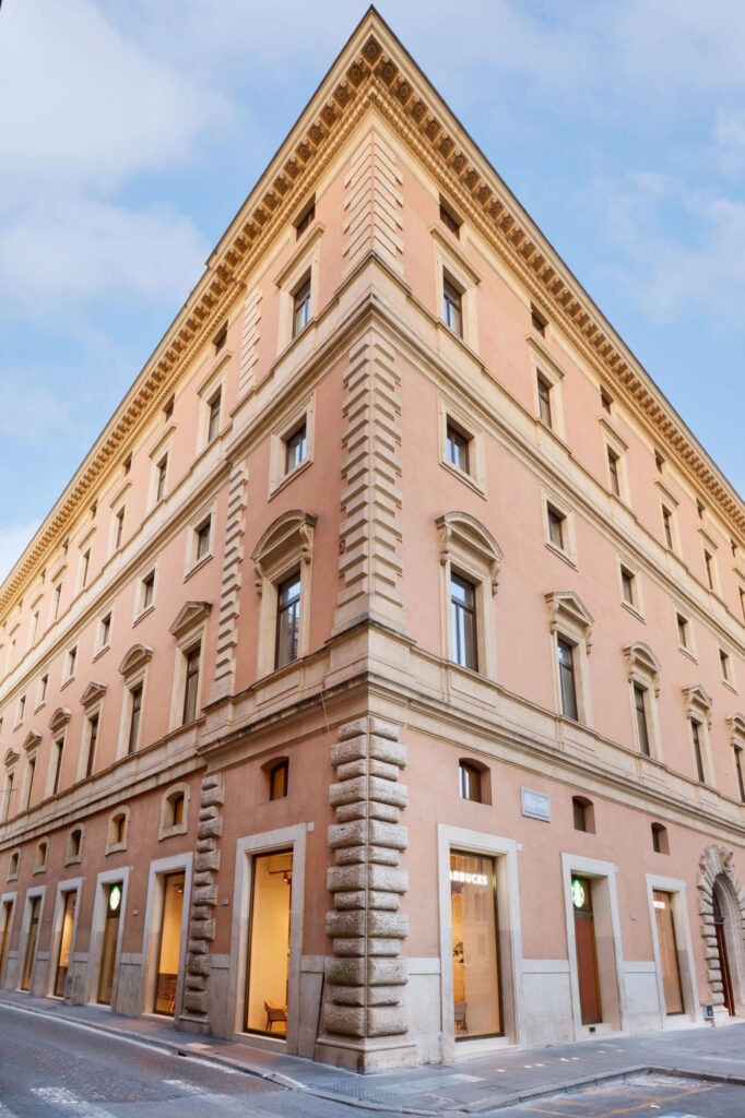 A corner view of a historic building with a pink facade, featuring decorative stonework and multiple windows. Two storefronts are visible, one displaying a Starbucks logo. The sky is clear with soft clouds.