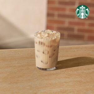 A clear glass filled with iced coffee, featuring ice cubes and a creamy, light brown color, sits on a wooden table.