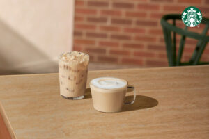 A glass of iced coffee with ice cubes sits next to a cup of creamy latte topped with froth, both on a wooden table.