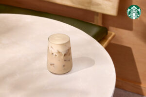 A glass of iced coffee sits on a round, light-colored table. The drink features a creamy top and visible ice cubes. A green bench is partially visible in the background.