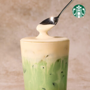 A tall glass filled with layered green matcha and creamy beige liquid, topped with a glossy layer. A spoon hovers above, dripping.