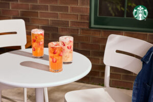 Three tall glasses of colorful drinks with ice and strawberries sit on a round white table, next to a white chair and a brick wall.
